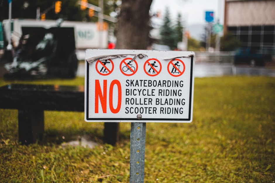 Close-up of a rectangular outdoor sign mounted on a metal post, displaying restrictions in red and black text that prohibit skateboarding, bicycle riding, roller blading, and scooter riding. The sign is situated on a grassy area next to a pavement, with a blurred background of trees, a street with vehicles including a black van, building structures, and traffic lights. The sign emphasizes the word 'NO' in large red letters with detailed service relevance to house removals and furniture transport by Man with Van Elm Park, supporting safe and compliant home relocation and moving logistics in Elm Park.