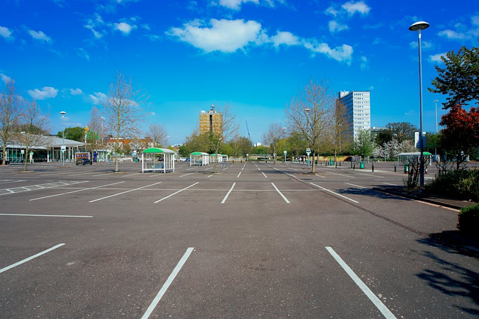A nearly empty parking lot with multiple parking bays marked by white lines on dark asphalt, situated in Elm Park under a bright blue sky with scattered clouds. Several leafless trees and some bushes are visible surrounding the area, along with small covered stalls or kiosks with green canopies on the left side. In the background, there are tall residential and office buildings, including a brown, multi-storey apartment block and a white high-rise, indicating an urban setting. The parking lot is illuminated by tall streetlights, one prominently visible on the right, and appears to be part of a larger retail or public area. The absence of moving vehicles and the orderly arrangement of parking spaces suggest a quiet, well-maintained environment suitable for home relocation or furniture transport activities, as managed by companies such as Man with Van Elm Park. This setting supports careful logistics and loading processes during packing and moving operations, especially given the accessible pavement and open space ideal for loading household items.