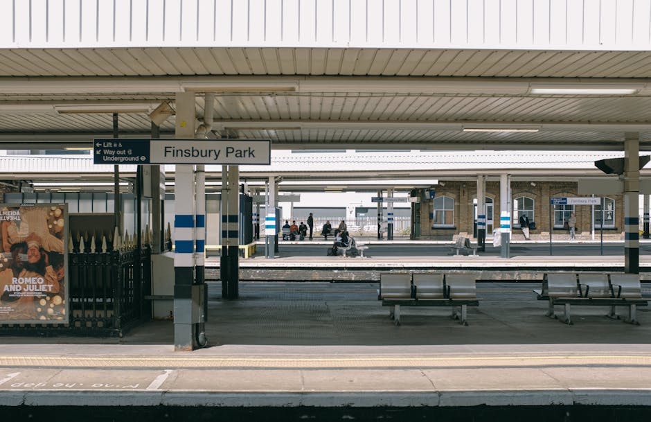 Wide view of Finsbury Park railway station platform during daytime, showing a covered waiting area with metal benches and blue-and-white striped pole markings. Several passengers are seated and standing on the platform, some using mobile devices. In the foreground, a black metal fence encloses an area with an advertisement poster for 'Romeo and Juliet' theatre show. The station sign reading 'Finsbury Park' is suspended from the ceiling structure. Behind the platform, multiple train tracks run parallel, with another platform and station building visible in the background, featuring brick walls and glass windows. The environment is well-lit with natural daylight, and the station appears clean and orderly, suitable for transportation and home relocation purposes. This scene reflects the typical setting where home relocation services like those offered by Man with Van Elm Park could load or unload furniture and packing materials, supporting efficient furniture transport and packing and moving logistics.