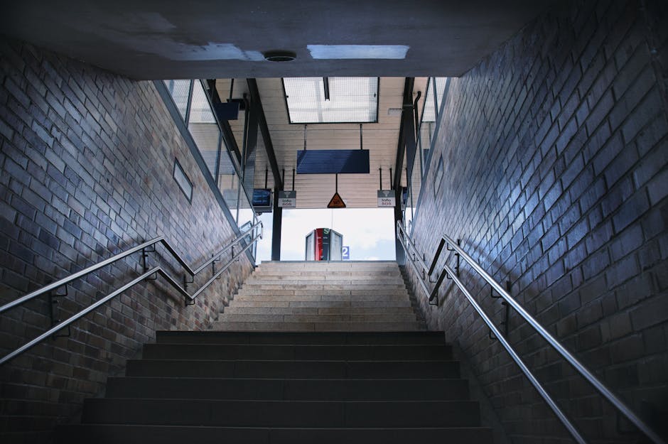 A set of indoor stairs leading up towards the exit of Elm Park station, featuring dark stairs with metal handrails on both sides. The walls on either side are constructed from exposed brick, with a window at the top allowing natural light to illuminate the stairway. Above the stairs, there are informational signs and a digital display. Through the open doorway at the top, part of the station platform and a telephone booth are visible outside. The image captures the staircase during daytime, emphasizing the structured environment typical of a train or metro station. Man with Van Elm Park may use such station locations during home relocation or furniture transport services, supporting efficient and accessible moving logistics.