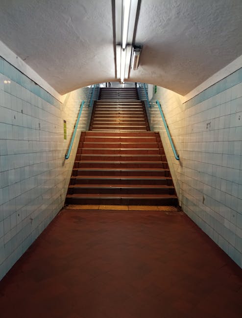 Inside a modern station with a high, glass-paneled roof allowing natural light to illuminate the space. The image shows a wide staircase with metallic handrails and steps leading upward, with several